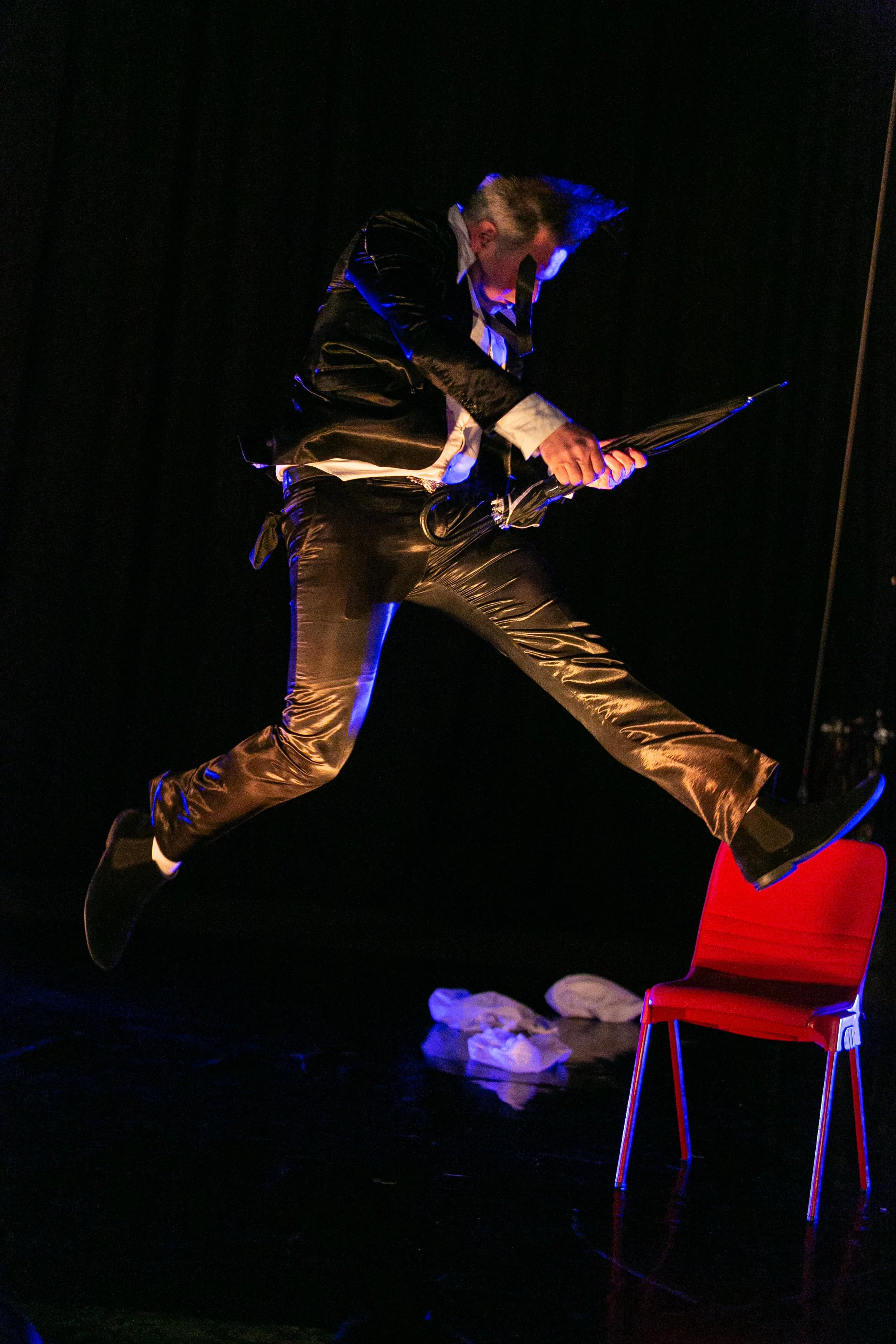 A performer in a shiny black suit and white shirt leaps over a red chair on a dark stage, holding a tablet or similar device.