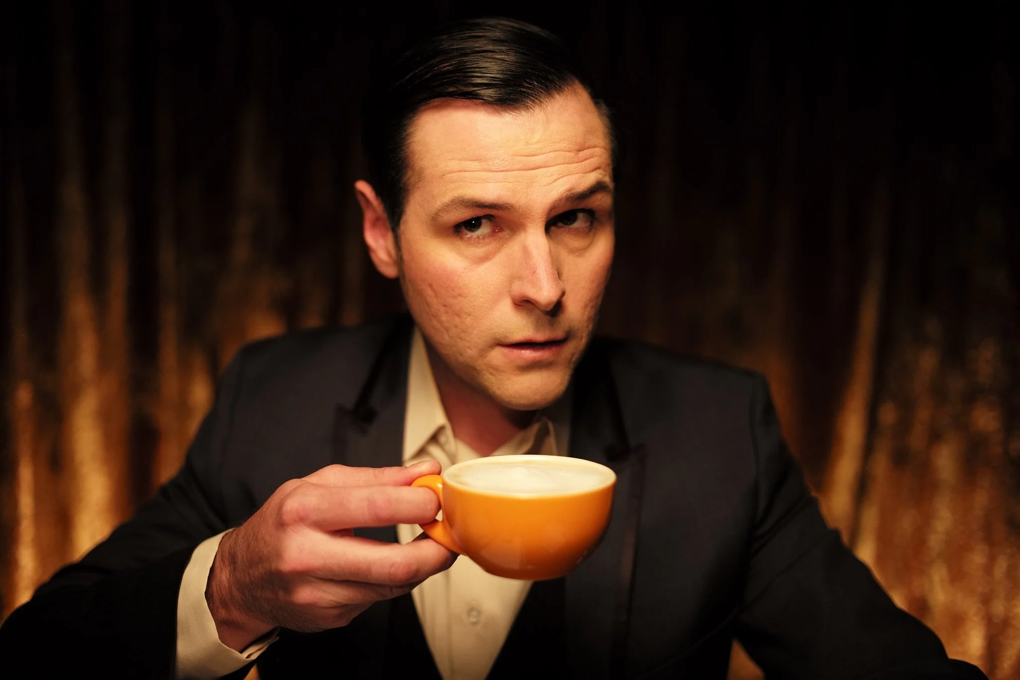A man with dark hair slicked back, wearing a black suit and cream shirt, holds an orange cup with a hot beverage, possibly coffee or tea, and looks at the camera against a dark, textured background.