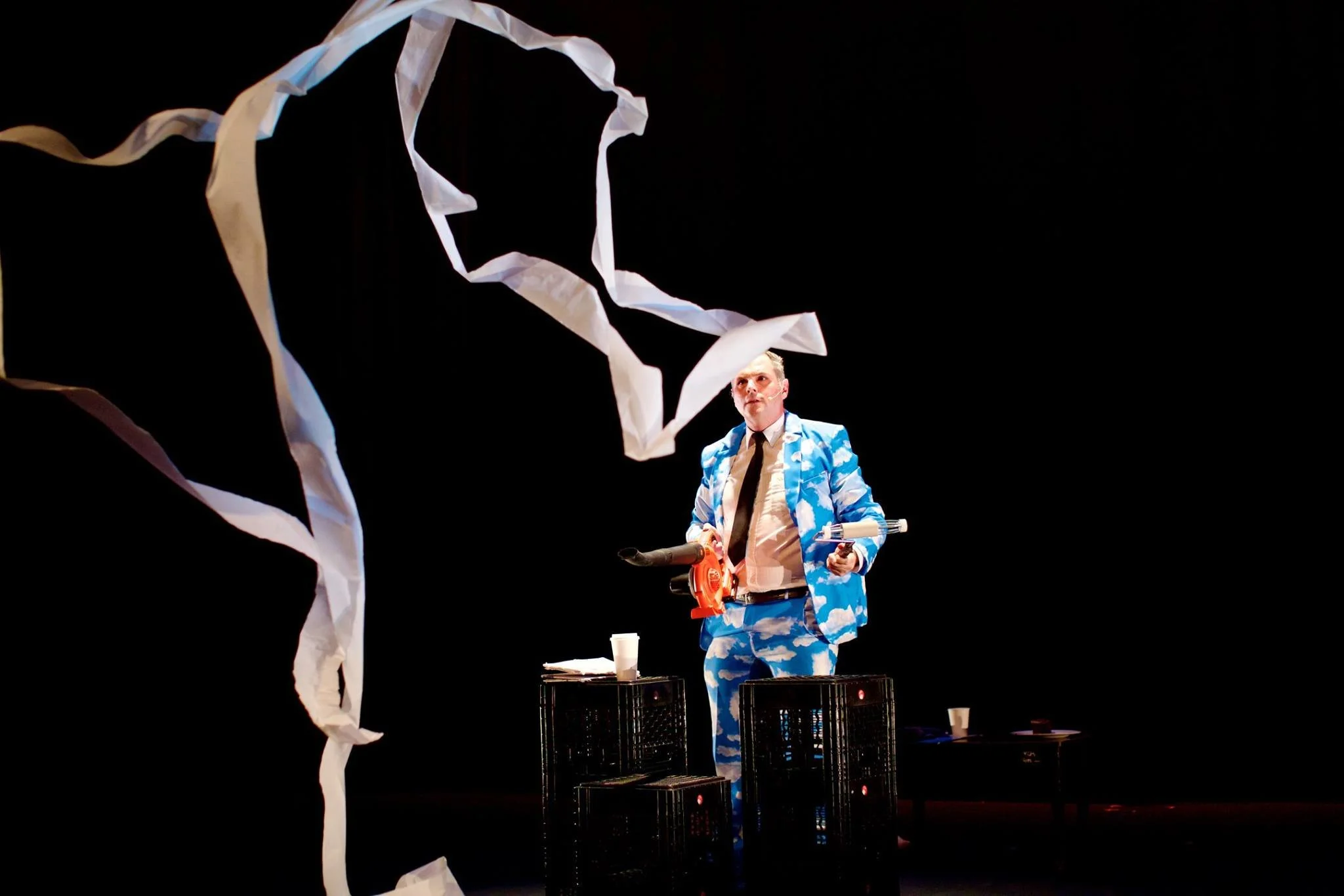 Performer standing on stage with torn white paper and black crates, wearing a blue and white cloud-patterned suit, holding a chainsaw and a gun, with a black background.