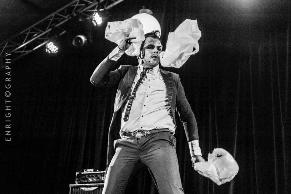 A male performer on stage, dressed in formal attire with a microphone headset, holding crumpled paper bags in both hands, with stage lights and curtain in the background.