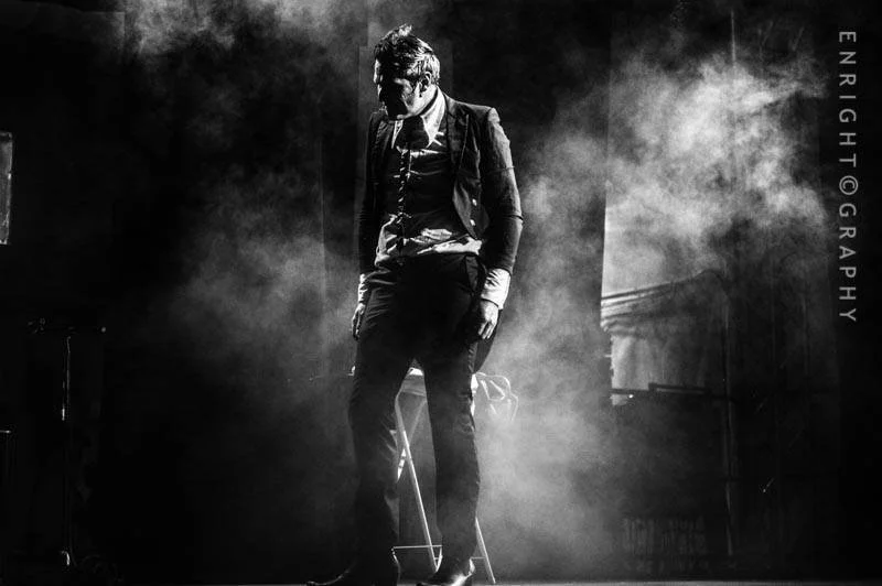 A man dressed in a business suit and backpack standing on stage surrounded by fog, with a contemplative pose, possibly during a performance or photo shoot.