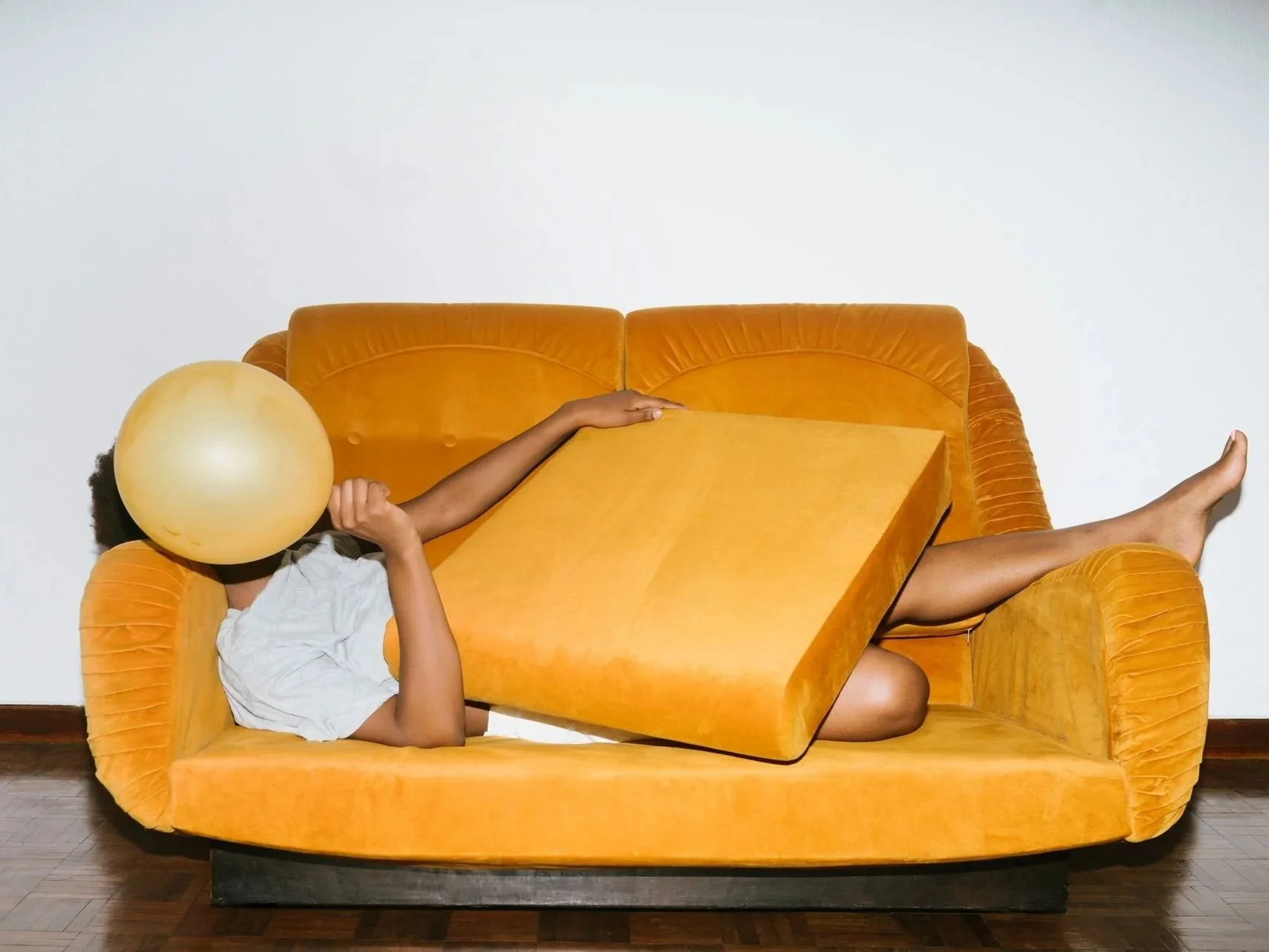 a person lies on a yellow sofa, holding a balloon over their face
