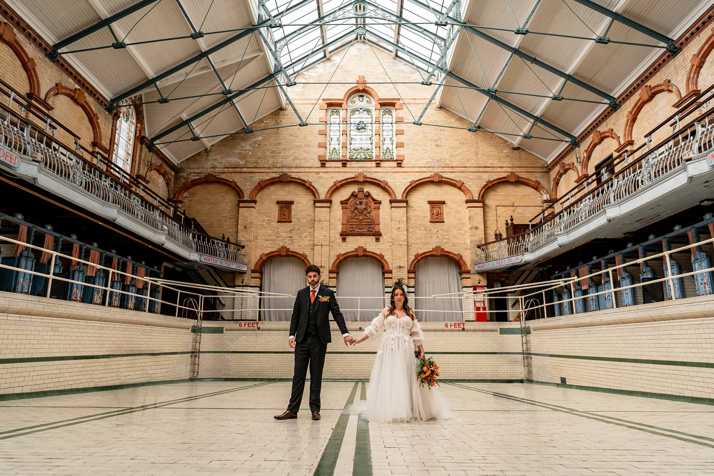 bride and groom holding hands looking at the camera in the empty swimming pool of Victoria Baths on their wedding day in manchester
