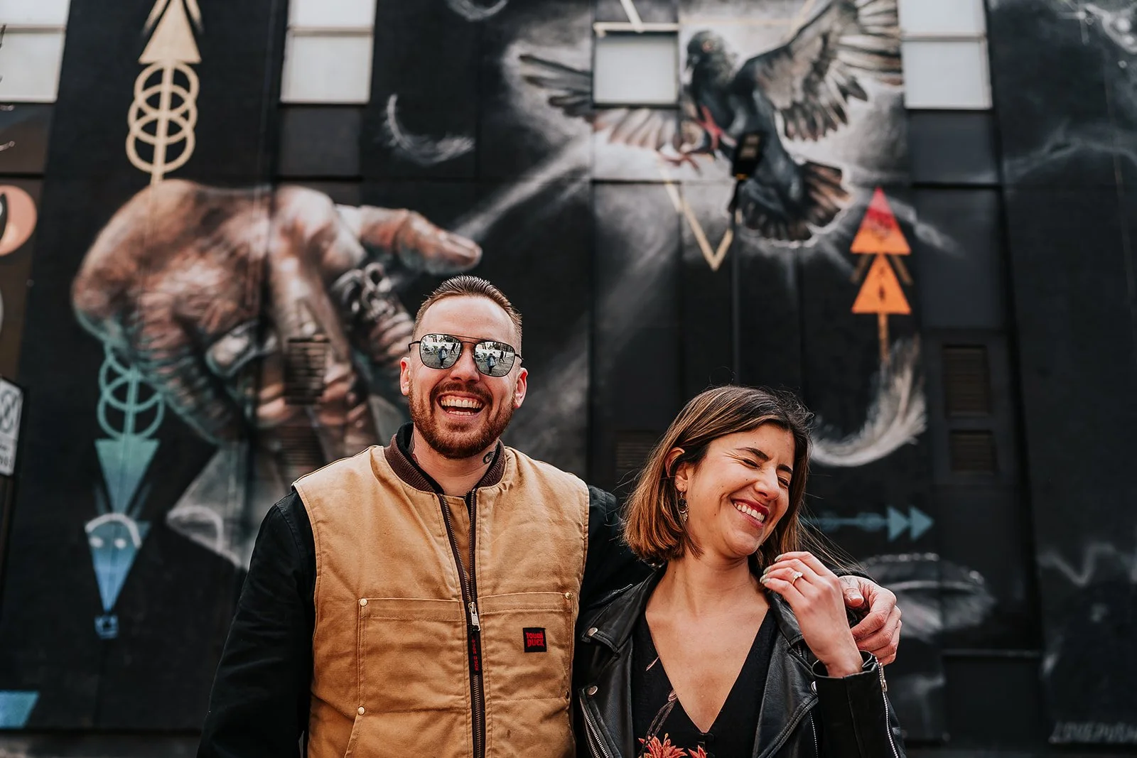 couple laughing at the camera in front of graffiti in London