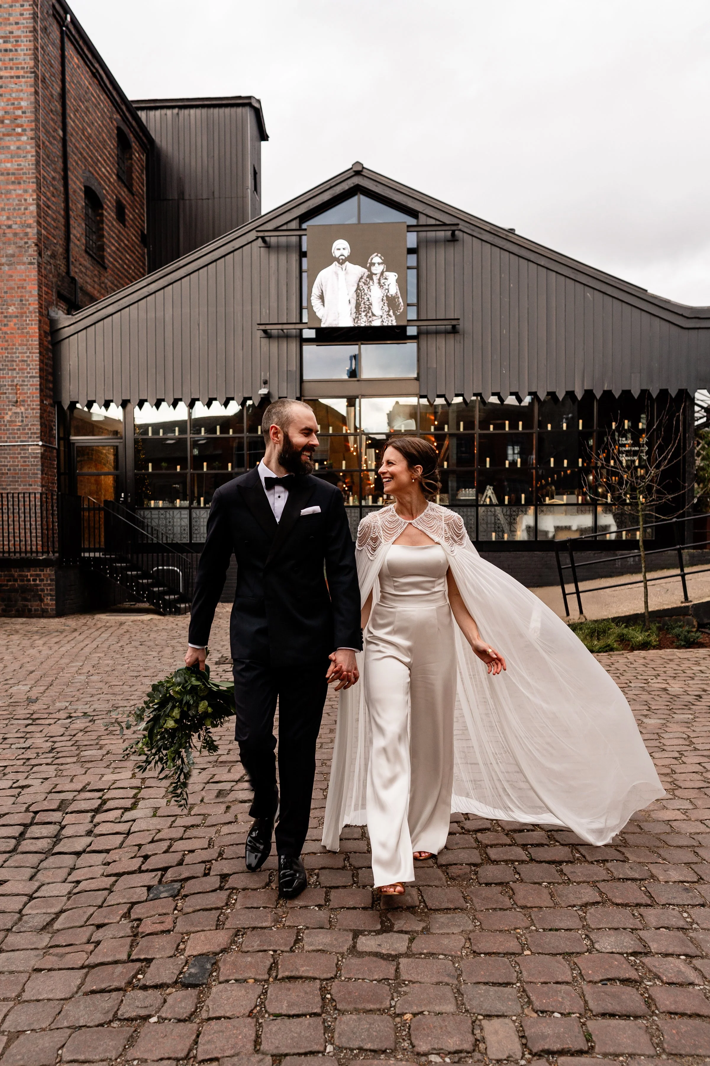 Bride and groom walking towards camera smiling at each other with their urban wedding venue in the back ground