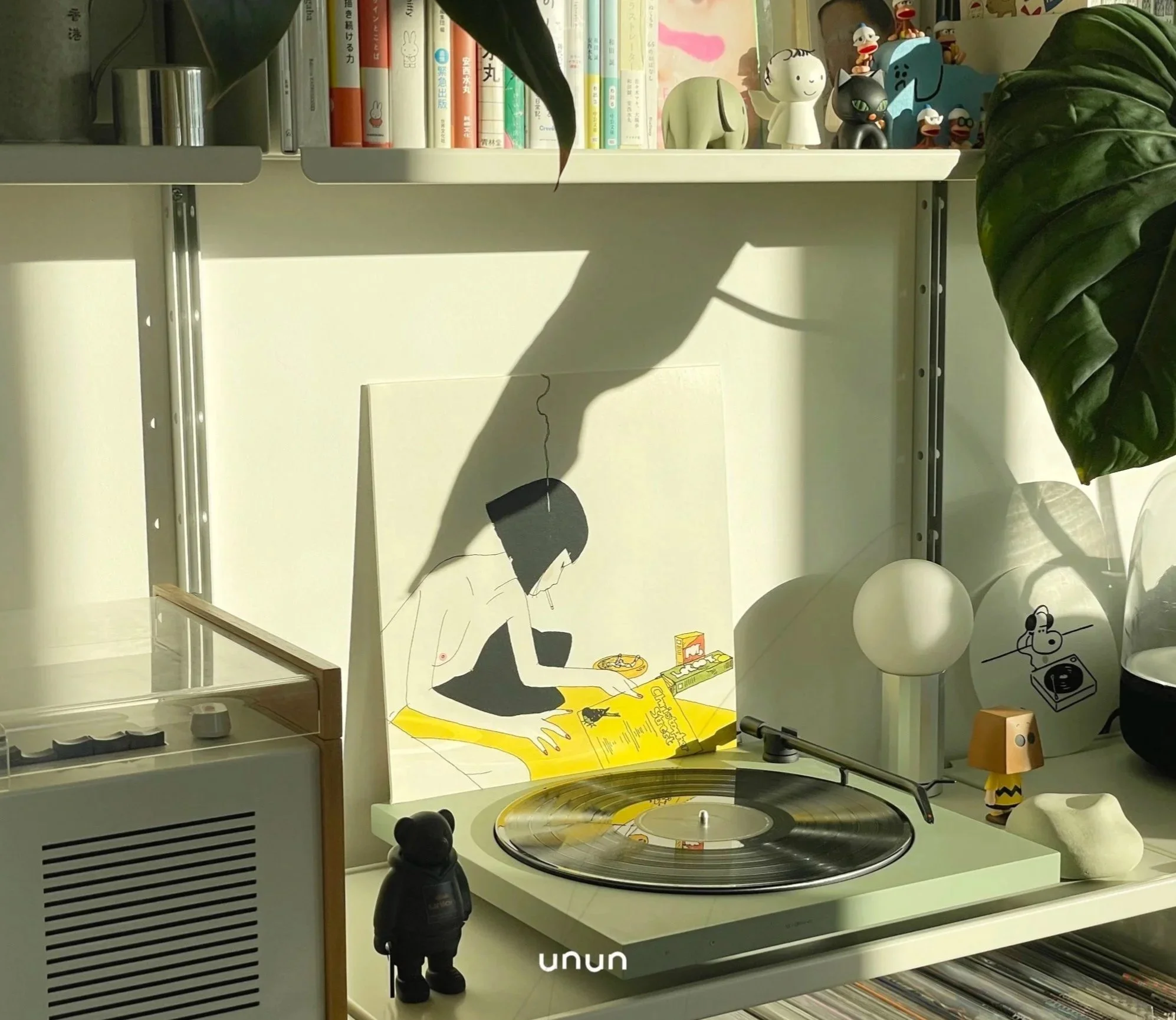 A Tone turntable with a vinyl record on it, decorated with various small figurines, a lamp, and a framed illustration of a woman reading a newspaper. There is a shelf with books and toys above, with sunlight casting shadows on the wall.