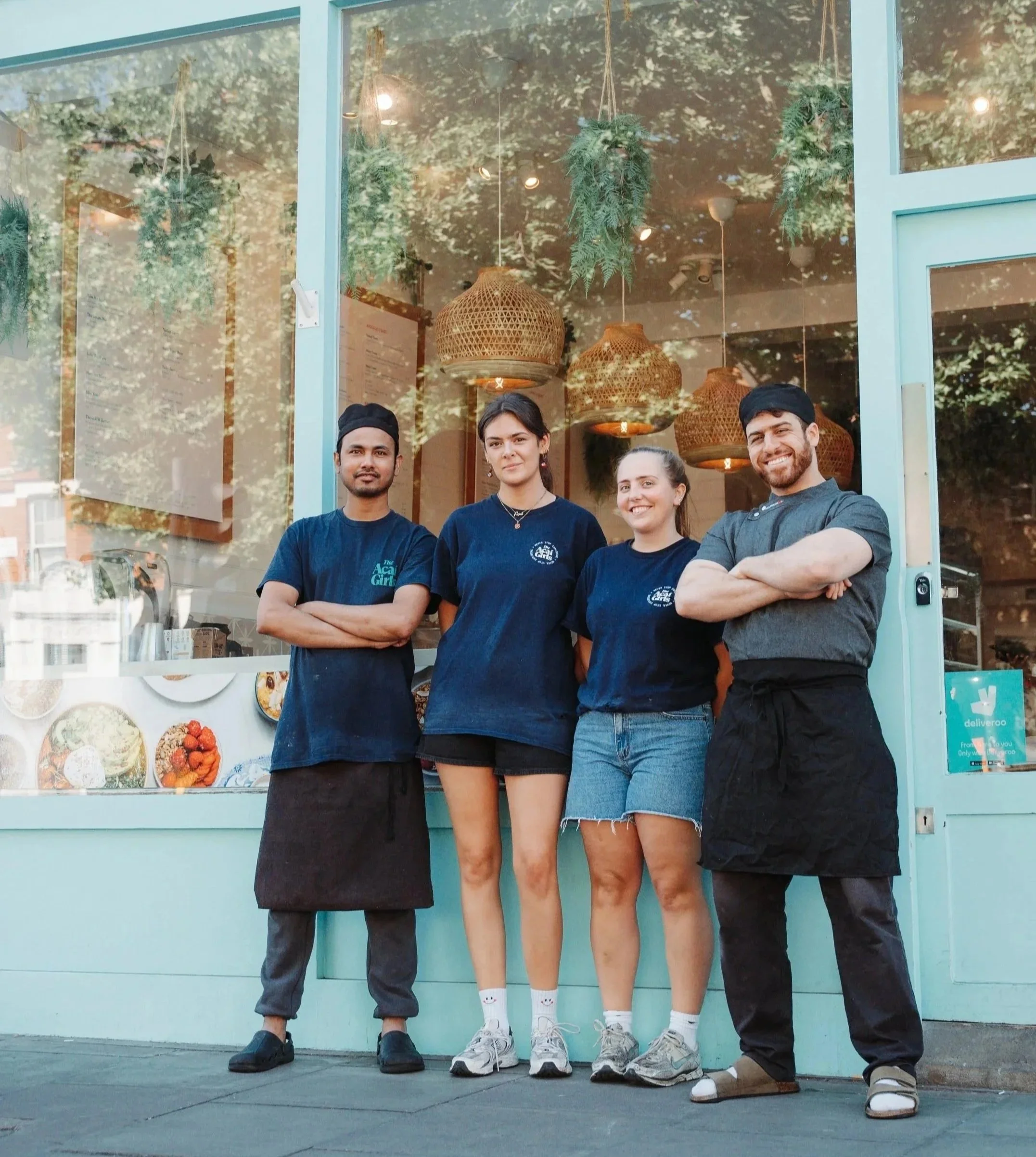 Four restaurant staff members standing outside a restaurant with large window, smiling, with hanging wicker lamps inside, and food photos displayed in the window.