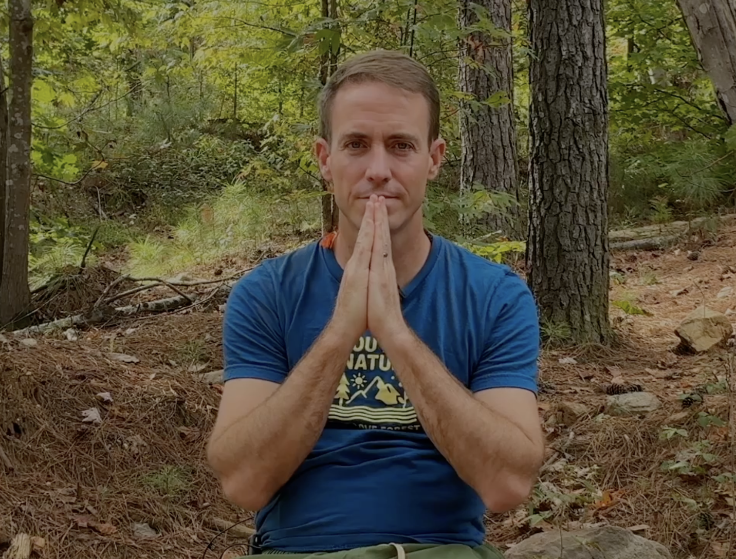 A man kneeling in a forest with his hands pressed together in a prayer position, surrounded by trees and foliage.