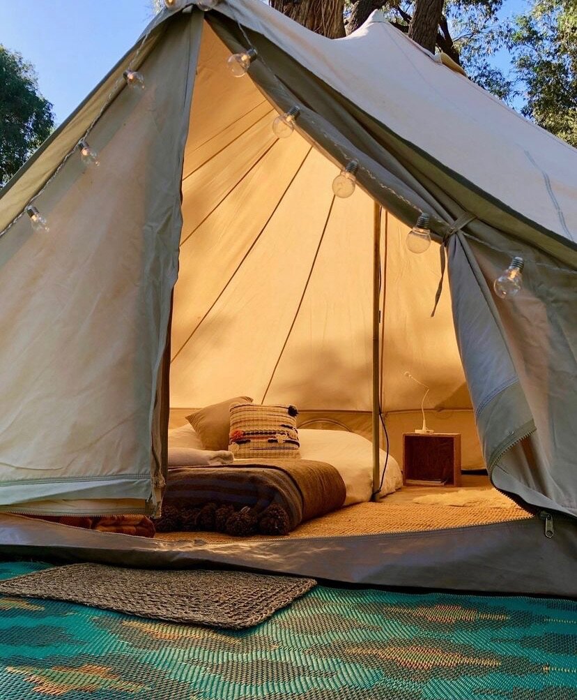 Inside a beige camping tent illuminated with string lights, showing a bed with pillows and blankets, a small wooden nightstand with a lamp, on a woven mat outside.