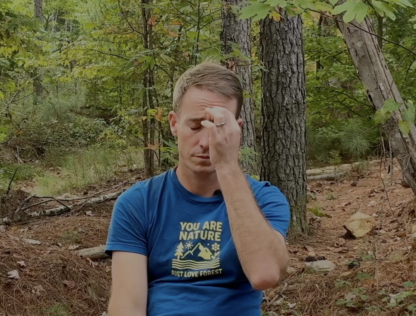 A man sits on the forest floor, rubbing his forehead with his hand, appearing tired or stressed, surrounded by trees and foliage.