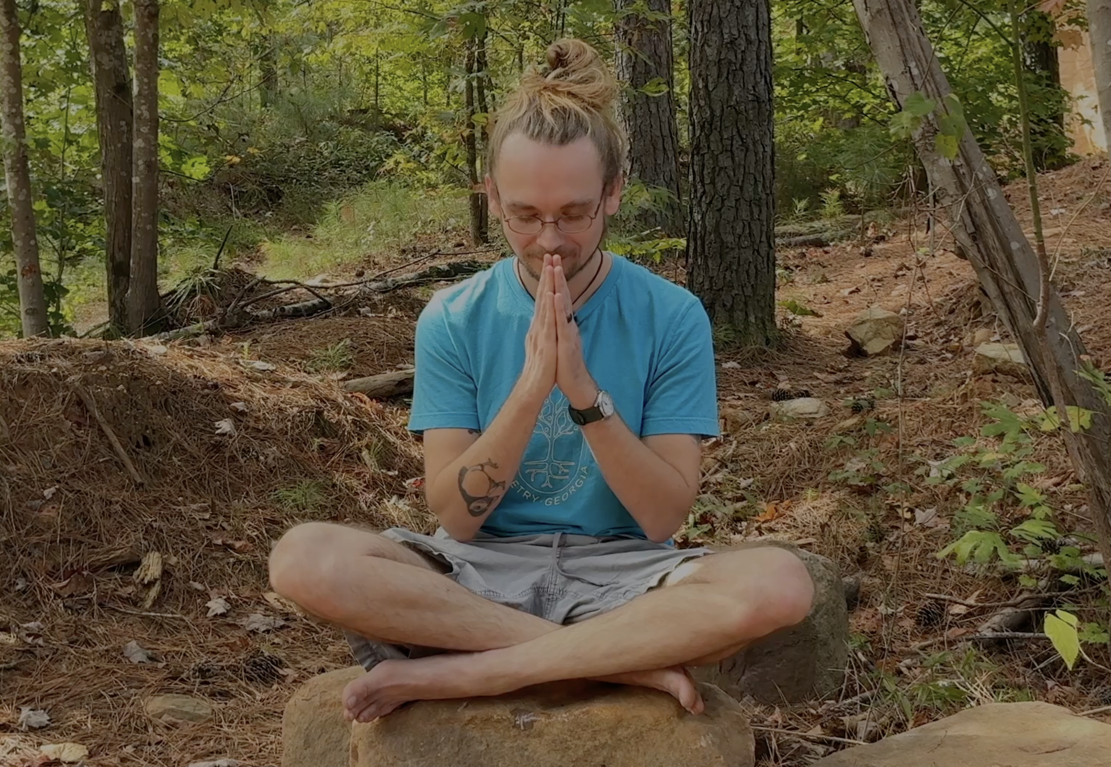 A man sitting cross-legged on a rock in a forest, with his hands pressed together in a prayer pose, eyes closed, and head bowed.