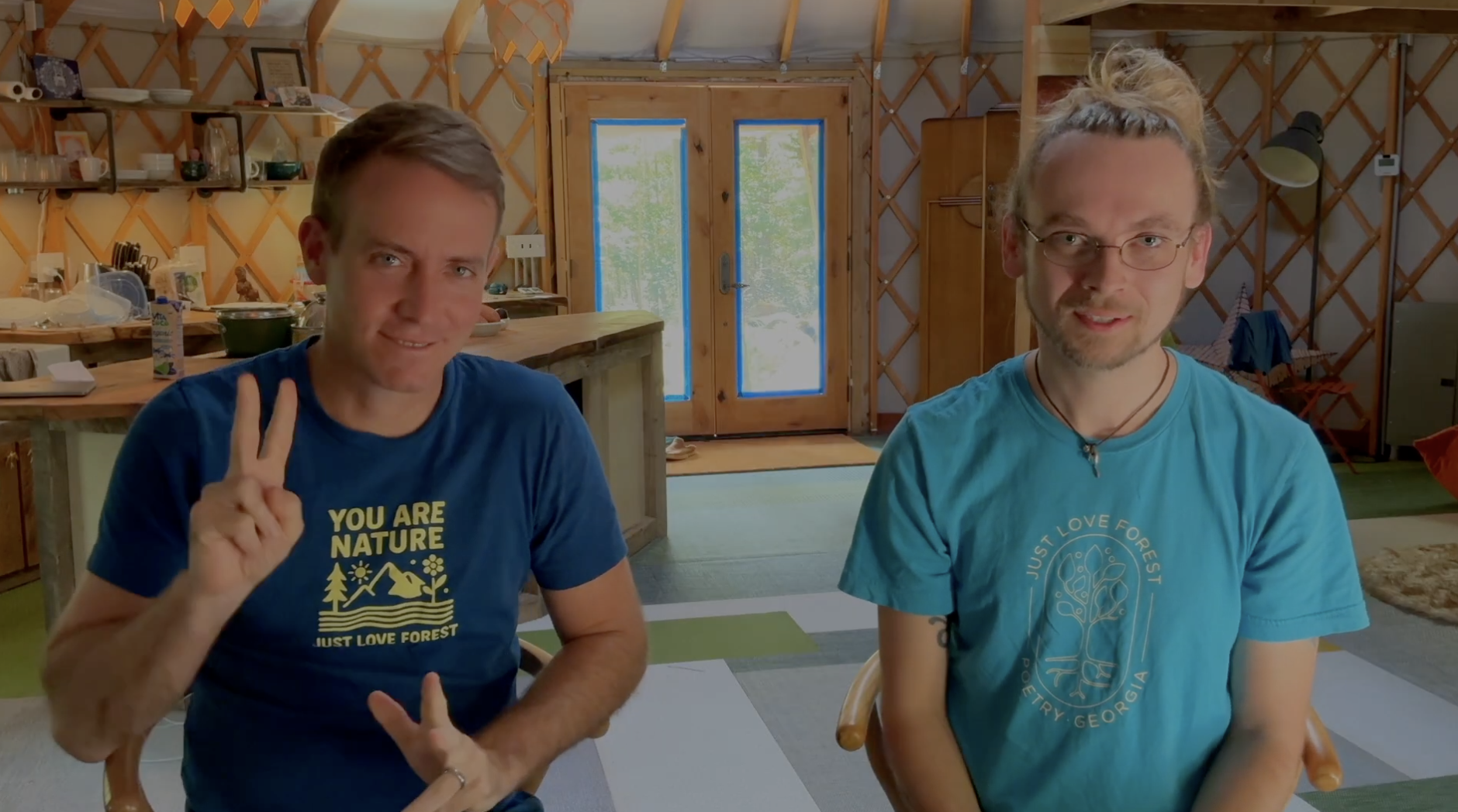 Two men sitting in a cozy, wooden yurt or cabin with lattice walls. One man on the left is making a peace sign and wearing a blue T-shirt with a nature-themed graphic. The other man on the right is wearing glasses and a blue T-shirt with a tree graphic. The background shows a kitchen area with shelves, dishes, and a patio door with blue trim leading outside into a wooded area.