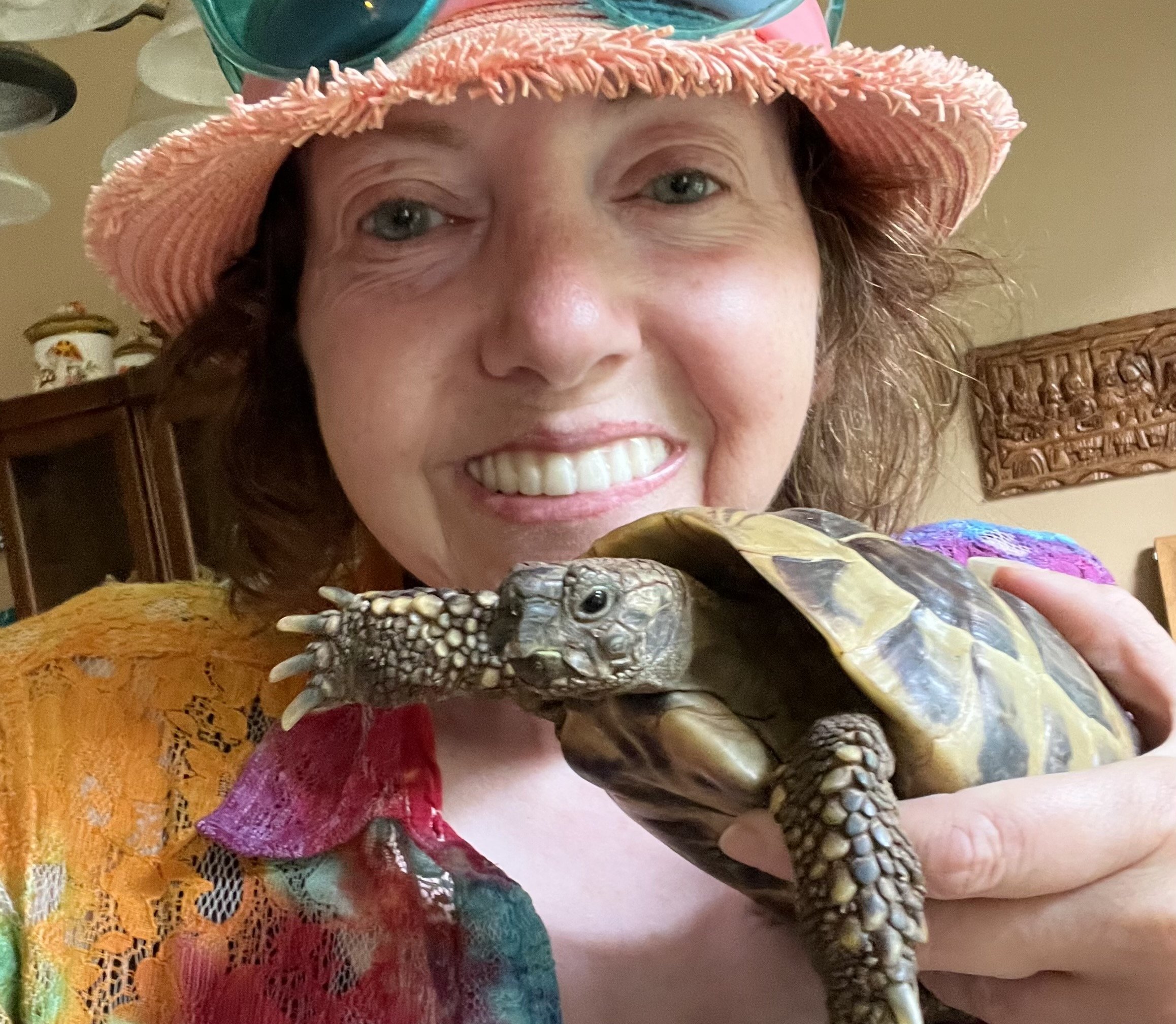 A woman smiling while holding a small turtle, wearing a pink sun hat and colorful clothing, in a room with wooden furniture and decorative items.