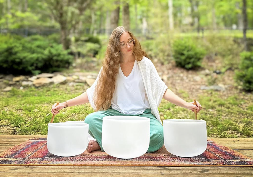 A woman with long curly hair, glasses, wearing a white top and green pants, sitting cross-legged on a rug outdoors, playing large white crystal singing bowls.