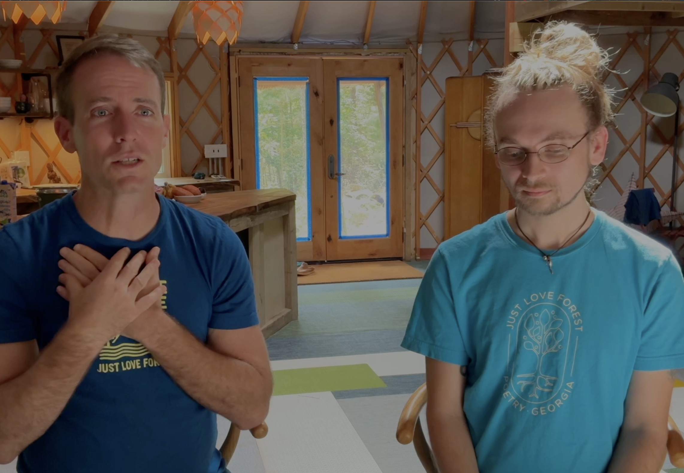 Two men sitting in a cozy, wooden interior with natural light coming through the windows and door. The man on the left, with short hair, is wearing a blue shirt and holding his hands over his chest. The man on the right, with glasses and long hair tied back, is wearing a turquoise shirt with a circular logo and text. The background features a wooden door with glass panels, a wooden cabinet, and a glimpse of greenery outside.