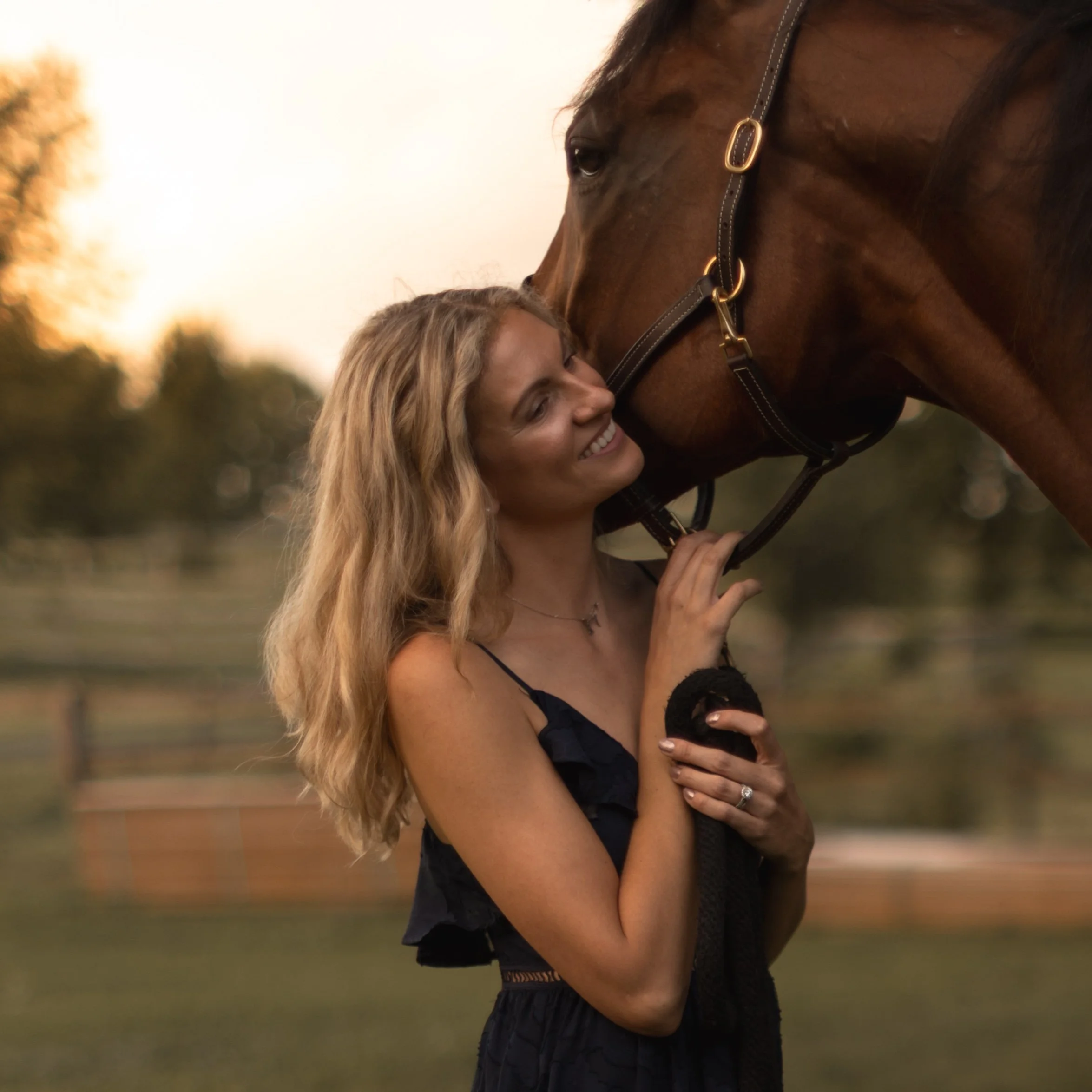 A woman with long blonde hair smiling and hugging a brown horse with a black saddle outdoors during sunset.