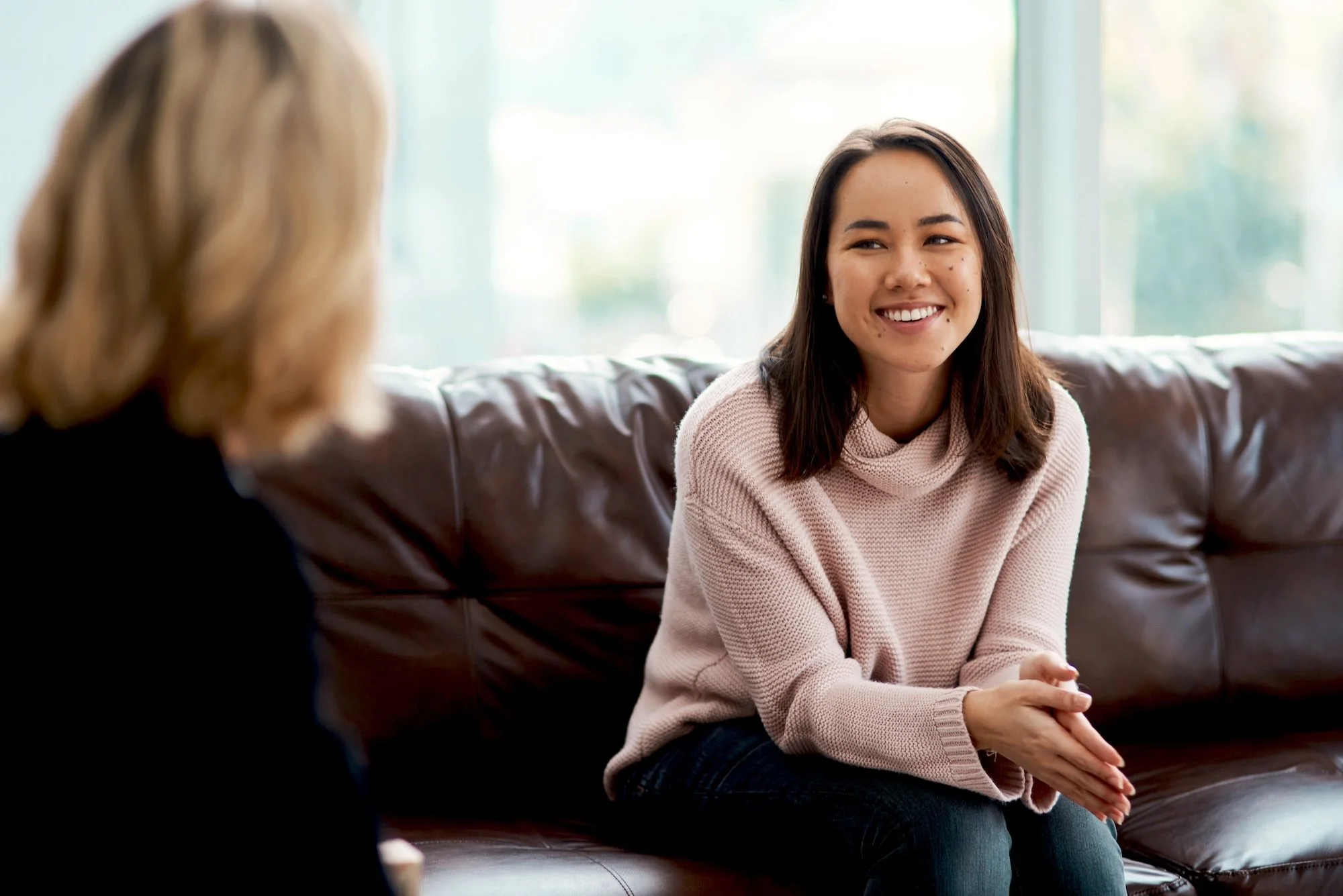 Two women sitting on a brown leather couch engaged in conversation, with a window in the background.