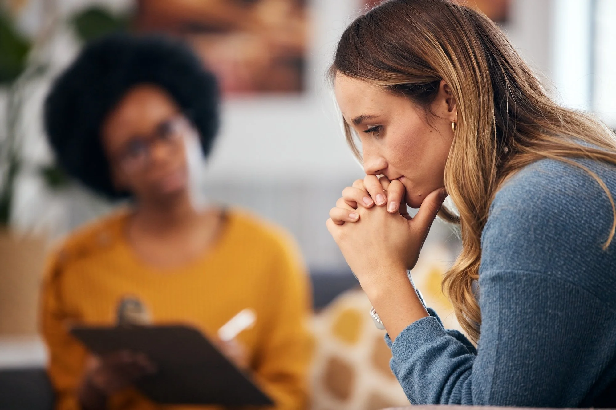 A woman with long light brown hair sitting with her hands clasped and head bowed, deep in thought or prayer, with a blurred person in a mustard-colored top in the background.