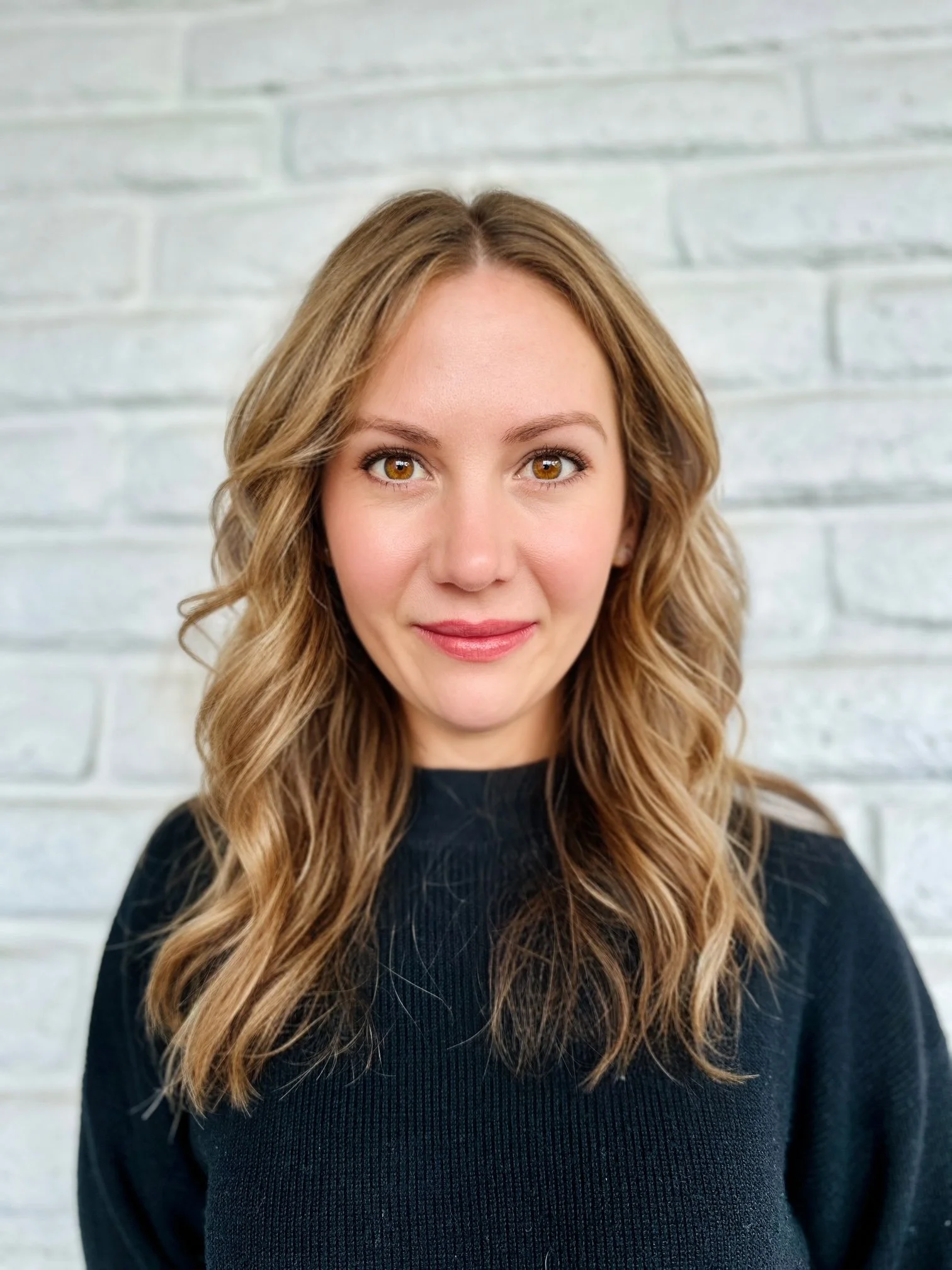 Close-up of a woman with wavy light brown hair, hazel eyes, wearing a black sweater, standing in front of a white brick wall.