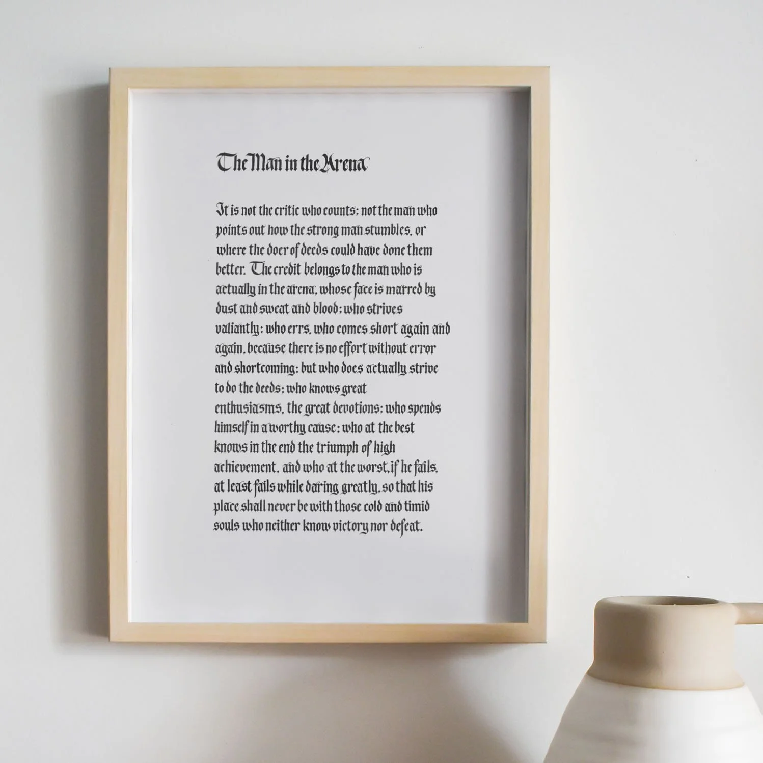 Framed quote titled 'The Man in the Arena' hanging on a wall, with a white ceramic vase partially visible in the lower right corner.