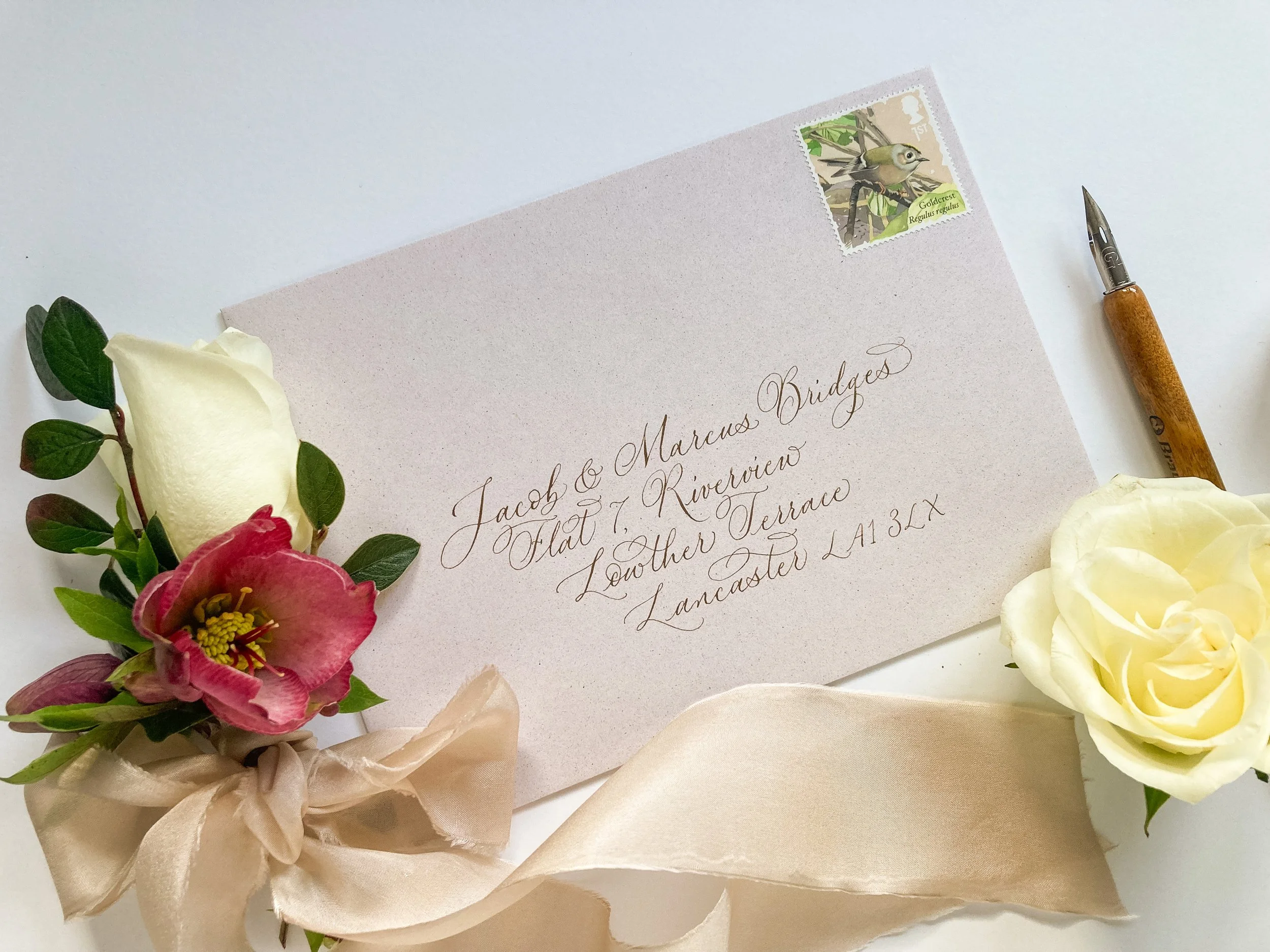 Pale recycled envelope with modern delicate handwritten calligraphy address, shown alongside a calligraphy dip pen, flowers and silk ribbon