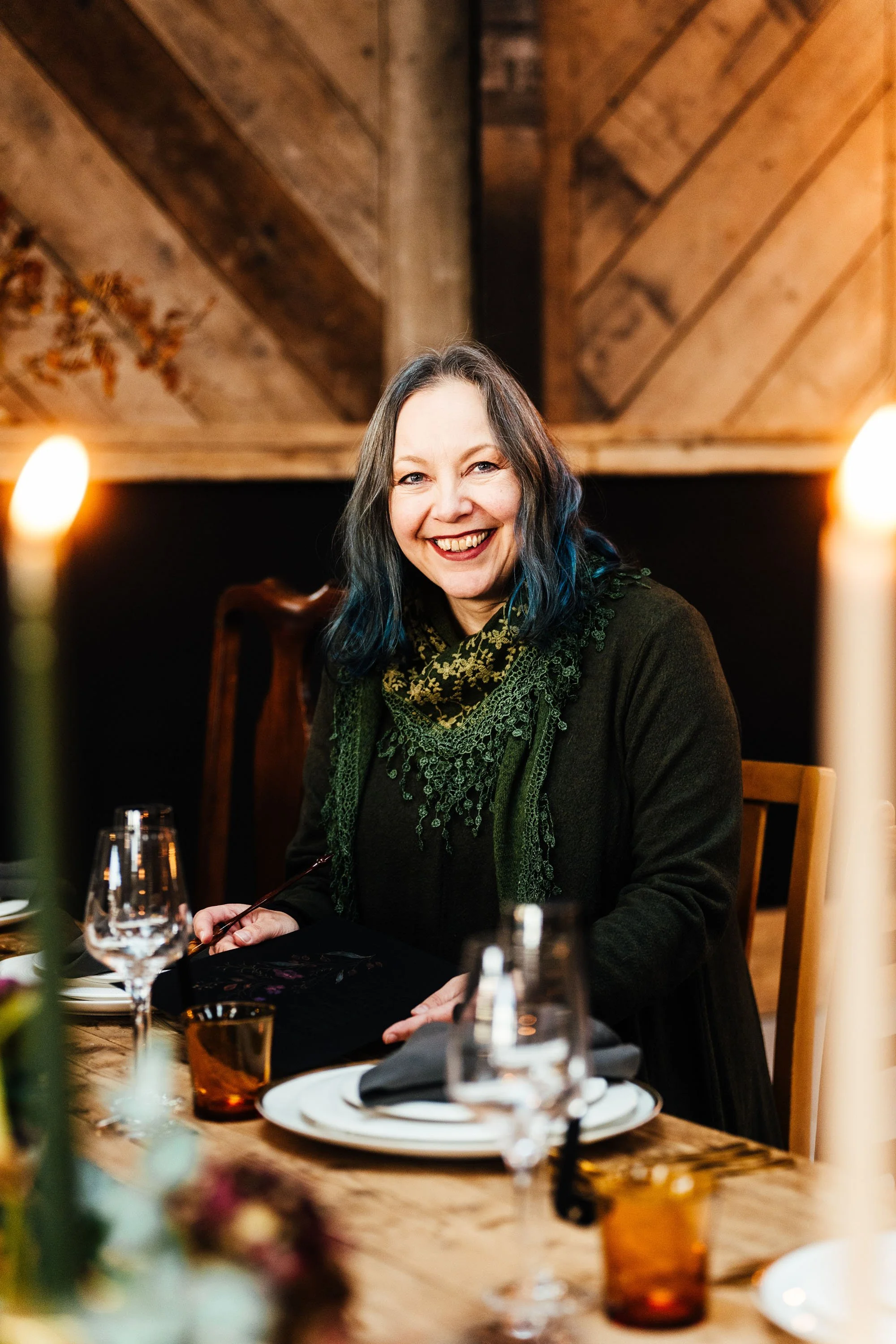 Calligrapher Claire Gould grinning at the camera, sat at a wedding table decorated with candles and amber glassware, with a calligraphy pen in her hand | Hannah Hall Photography at Ghyll Barn Cumbria