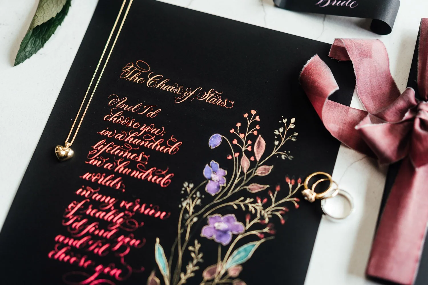 Calligraphy poem "The Chaos of Stars" in colour shift ink on black paper, with a botanical illustration. A gold heart necklace is on top of the poem, with silk ribbon to one side