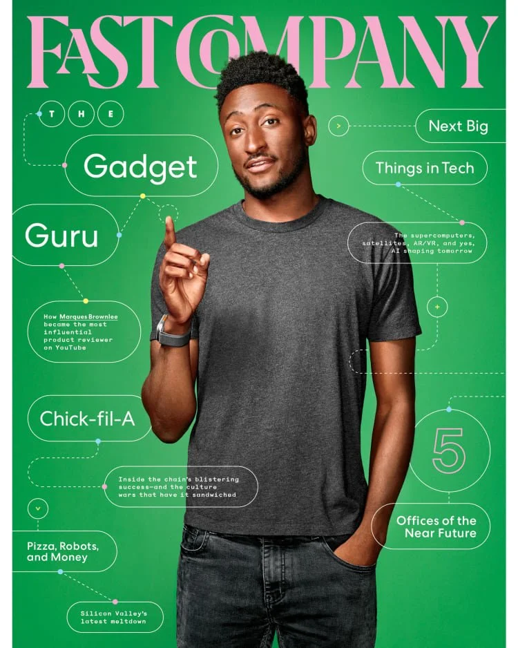 A young man with short curly hair and a beard wearing a gray t-shirt and black jeans, standing against a green background with tech-related headlines and graphics. He is pointing upwards with his right index finger.