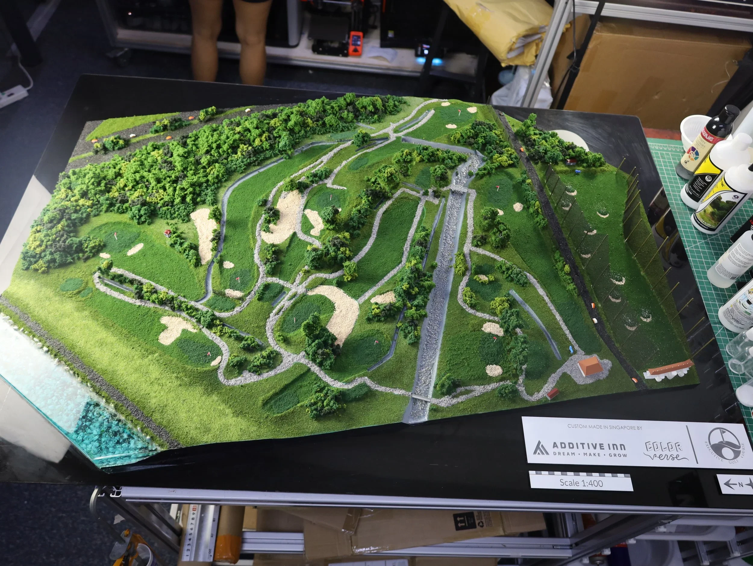 A detailed miniature model of a golf course with pathways, trees, sand traps, and grassy areas, displayed on a table with bottles of modeling supplies nearby.