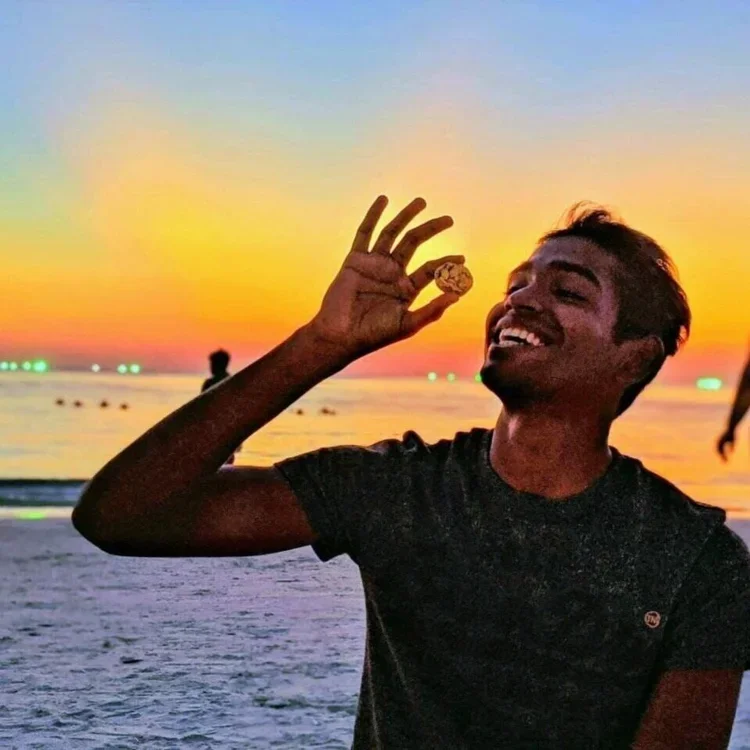 A man smiling and holding a small object, possibly a seashell or stone, near his face at the beach during sunset.