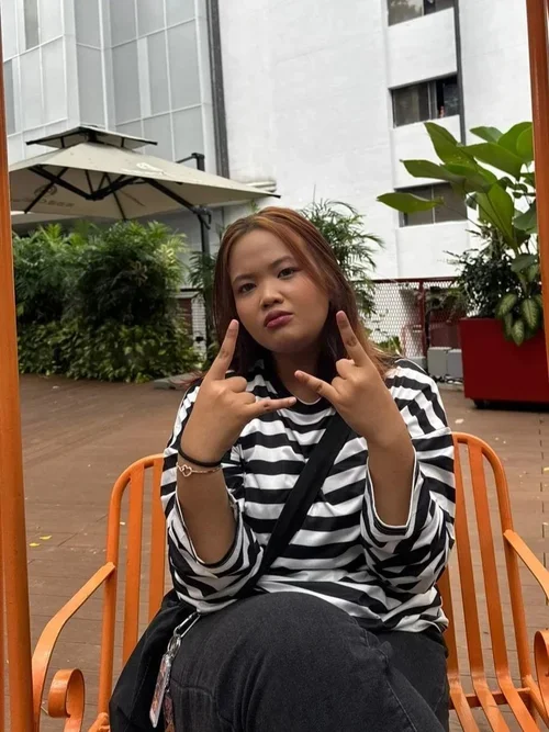 A young woman with light brown hair and a striped black and white shirt sitting outside on an orange metal chair, making a hand gesture with both hands, in an urban outdoor setting with greenery and modern buildings in the background.