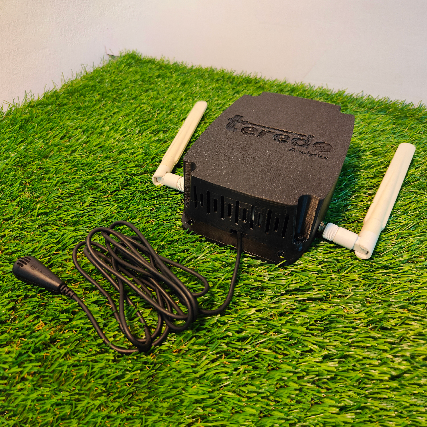 A black wireless router with two white antennas, a power cable, positioned on artificial grass.