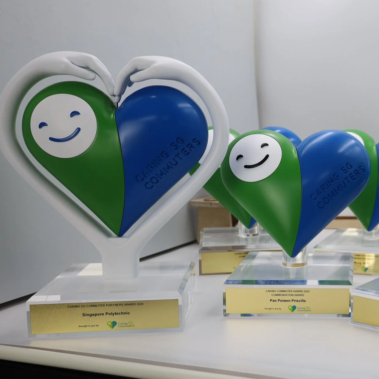 Two award trophies shaped like hearts with smiling faces and the text 'Caring SG Commuters'. One is larger with green and blue colors and a white frame, and the smaller one is similar in design. Both are displayed on a white surface.