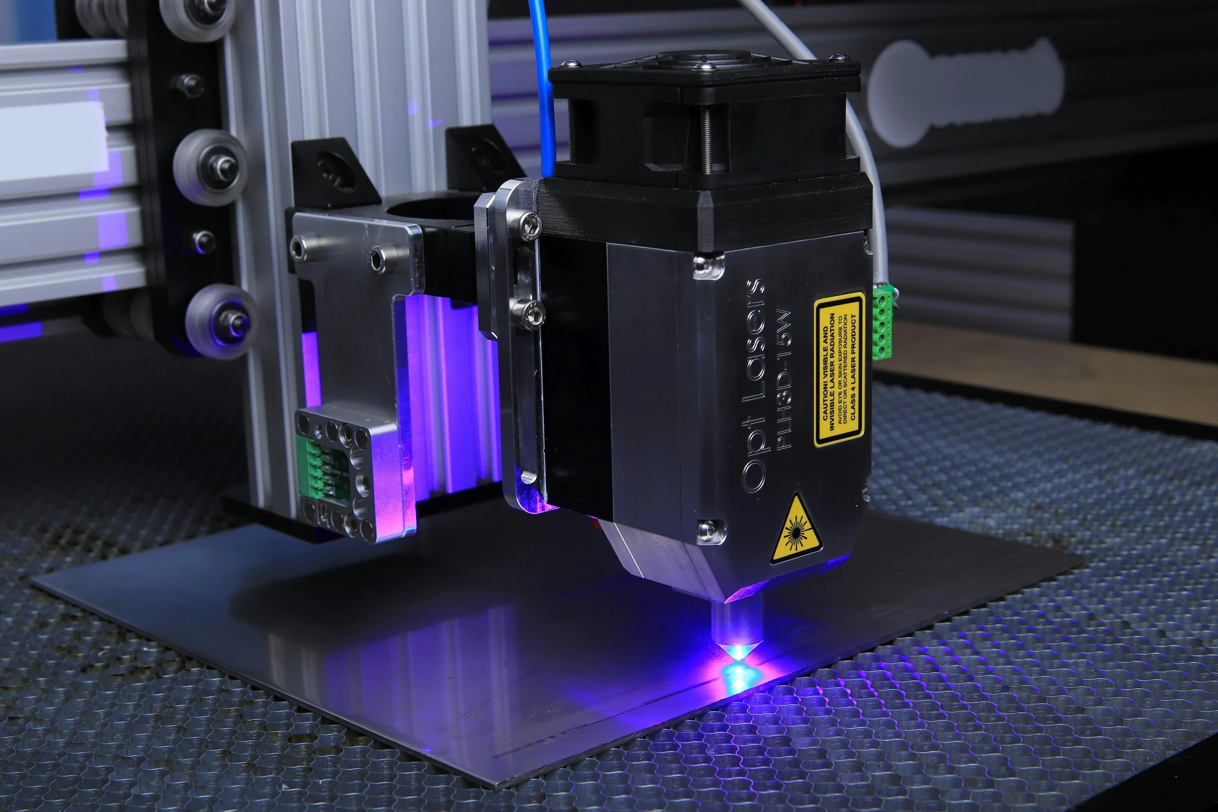 A laser engraving machine with a glowing blue laser beam working on metal, surrounded by an aluminum frame and electrical components.