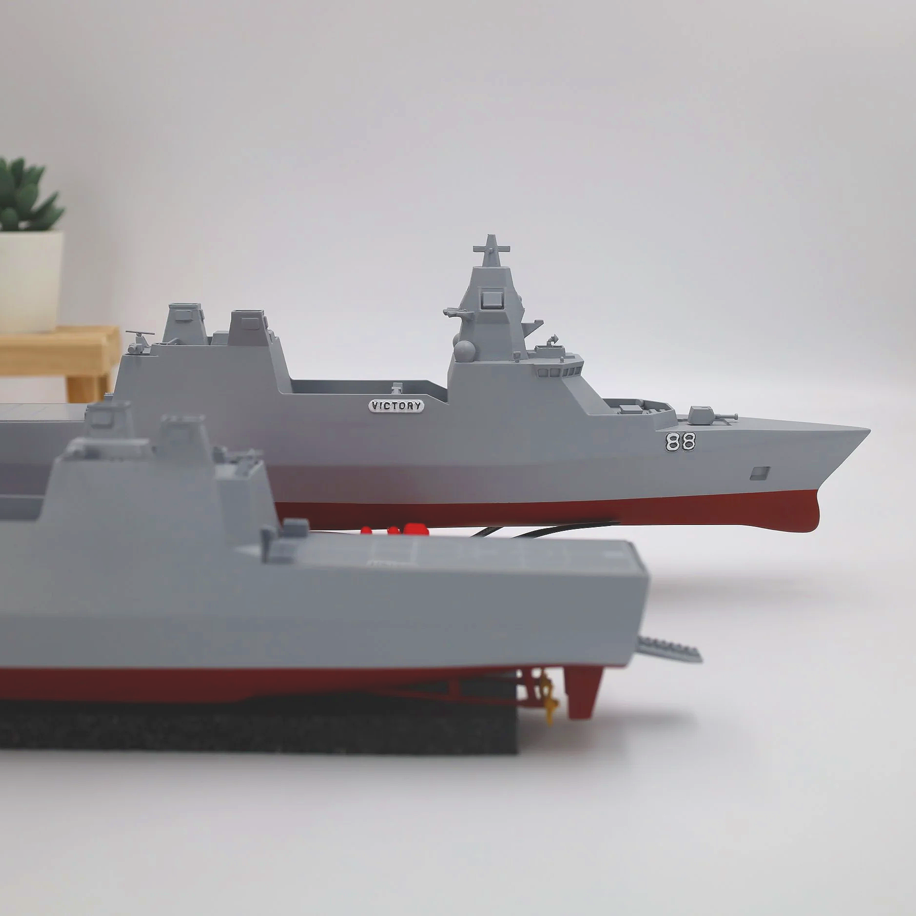 Model ships of different sizes in gray and red are placed on a white surface with a small wooden table and a potted plant in the background.