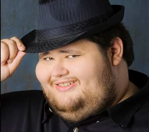 A man smiling, wearing a black shirt and a dark fedora hat, holding the brim of his hat with his right hand.