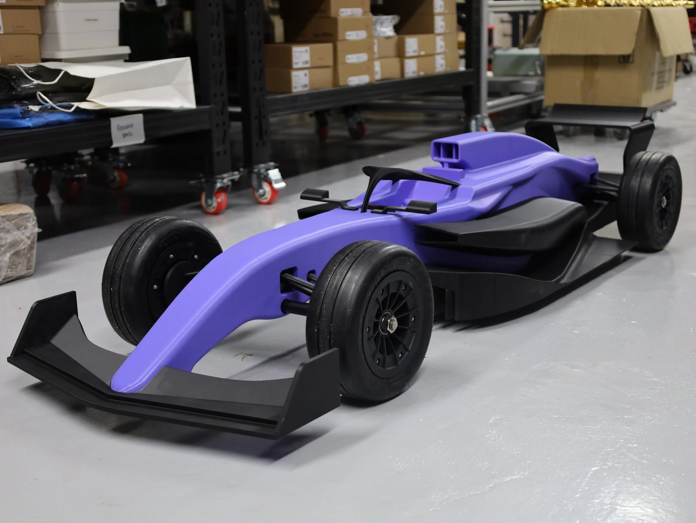 A purple and black toy race car with slick black tires, placed on a polished concrete floor in a workshop or storage area with metal shelves, boxes, and carts in the background.