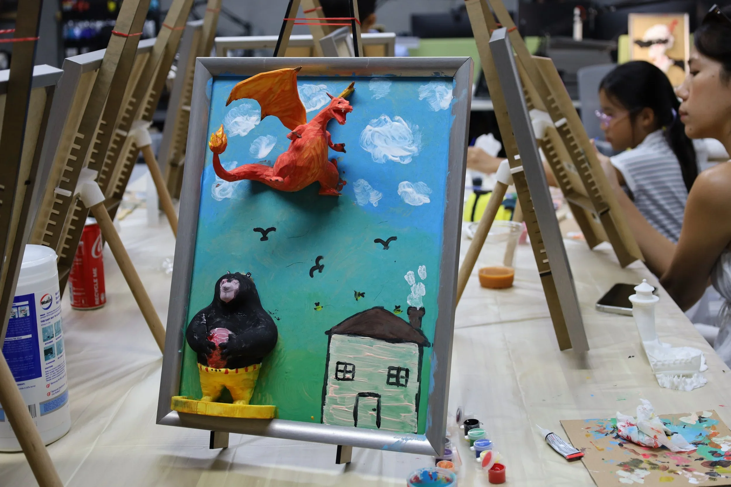 A 3D painted picture of a red dragon, a black gorilla with yellow pants, and a simple house with a chimney on a bright green grass field against a blue sky with white clouds, enjoyed by children painting at an art class.