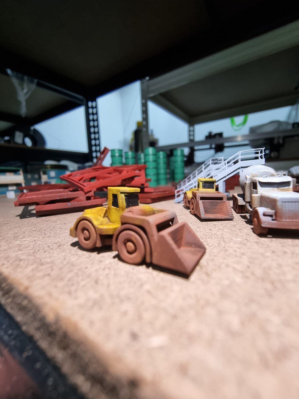 A collection of toy construction vehicles and equipment, including bulldozers and cranes, arranged on a workshop surface with storage shelves and green blocks in the background.
