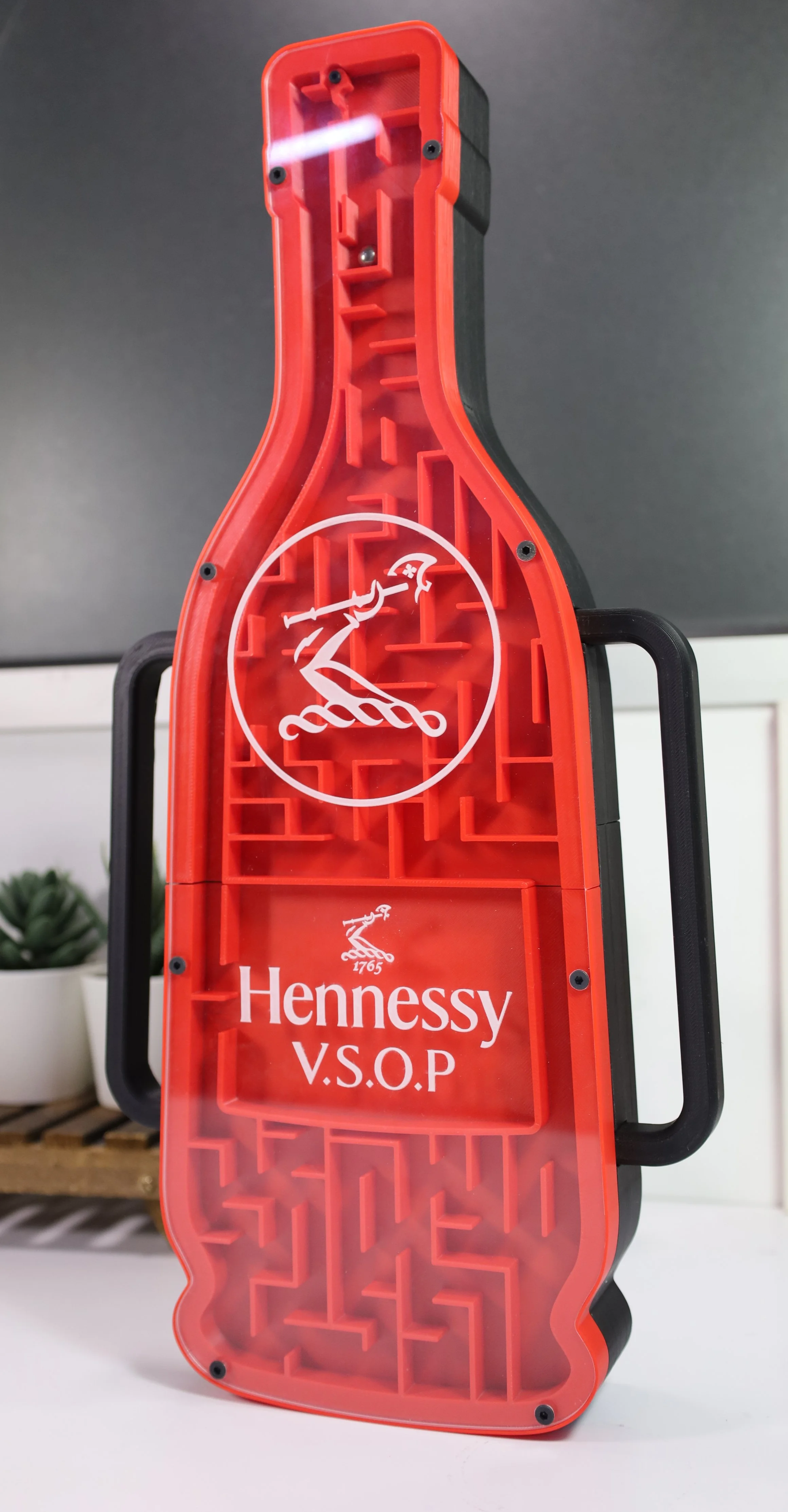 A red and black plastic case with the Hennessy V.S.O.P logo on its side, shaped like a shoe sole with internal compartments, stands upright on a white surface.