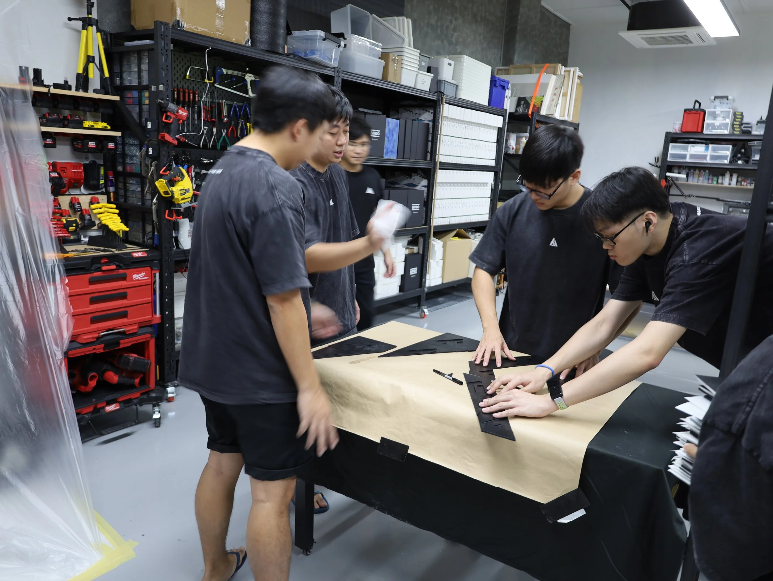 Five young men working together around a table in a workshop, sorting black material pieces, with tools and shelves of supplies behind them.