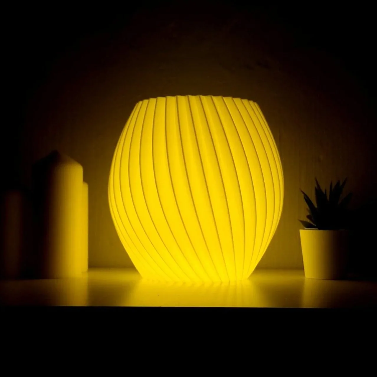 A glowing yellow contemporary table lamp with vertical slats, placed on a surface with two potted plants on either side, one tall cylindrical and one shorter with spiky leaves, in a dark room.