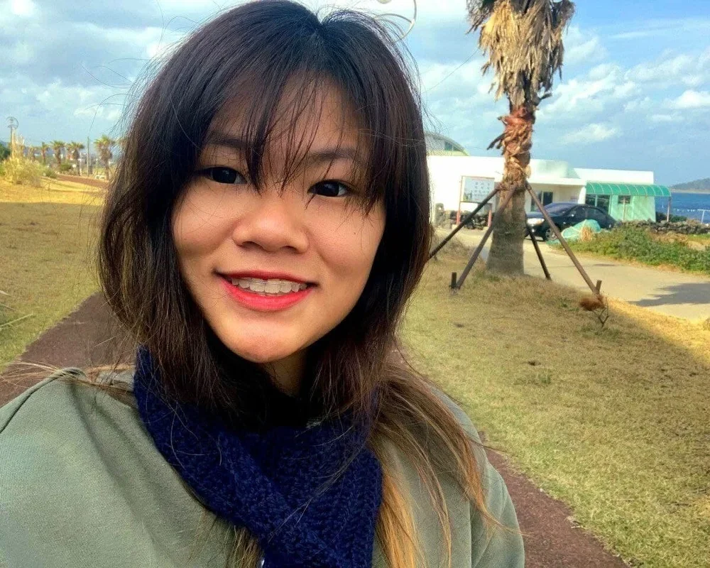 A woman smiling outdoors near a beach with palm trees, a building, and a parking lot in the background.