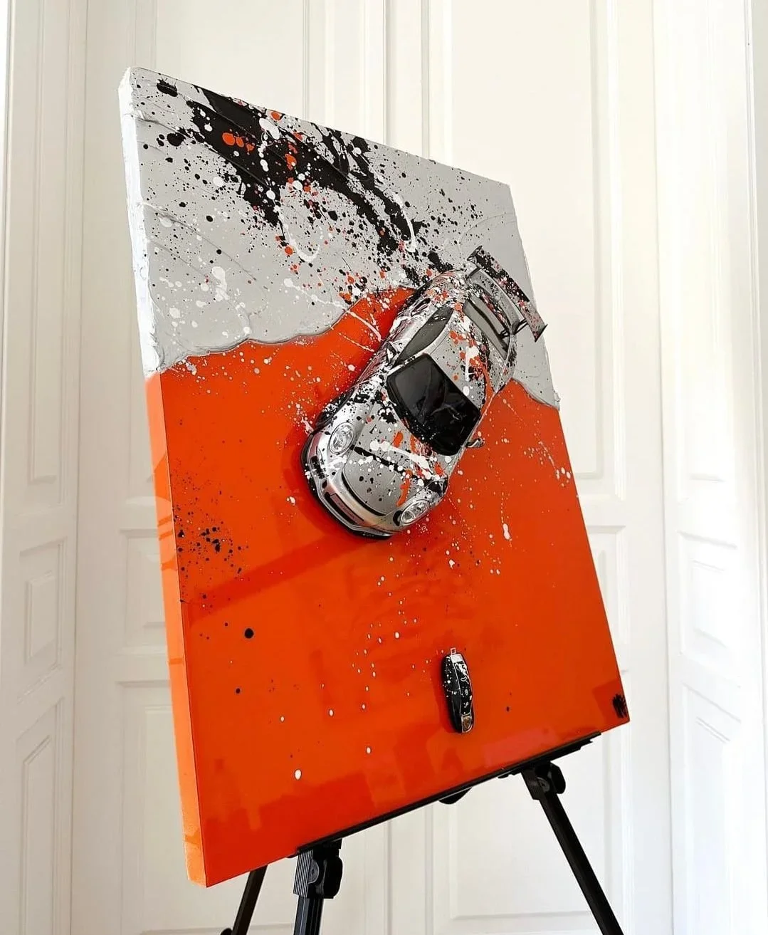 Mixed-media art piece with a car on an abstract background featuring splashes of black, orange, and white paint, mounted on a stand.