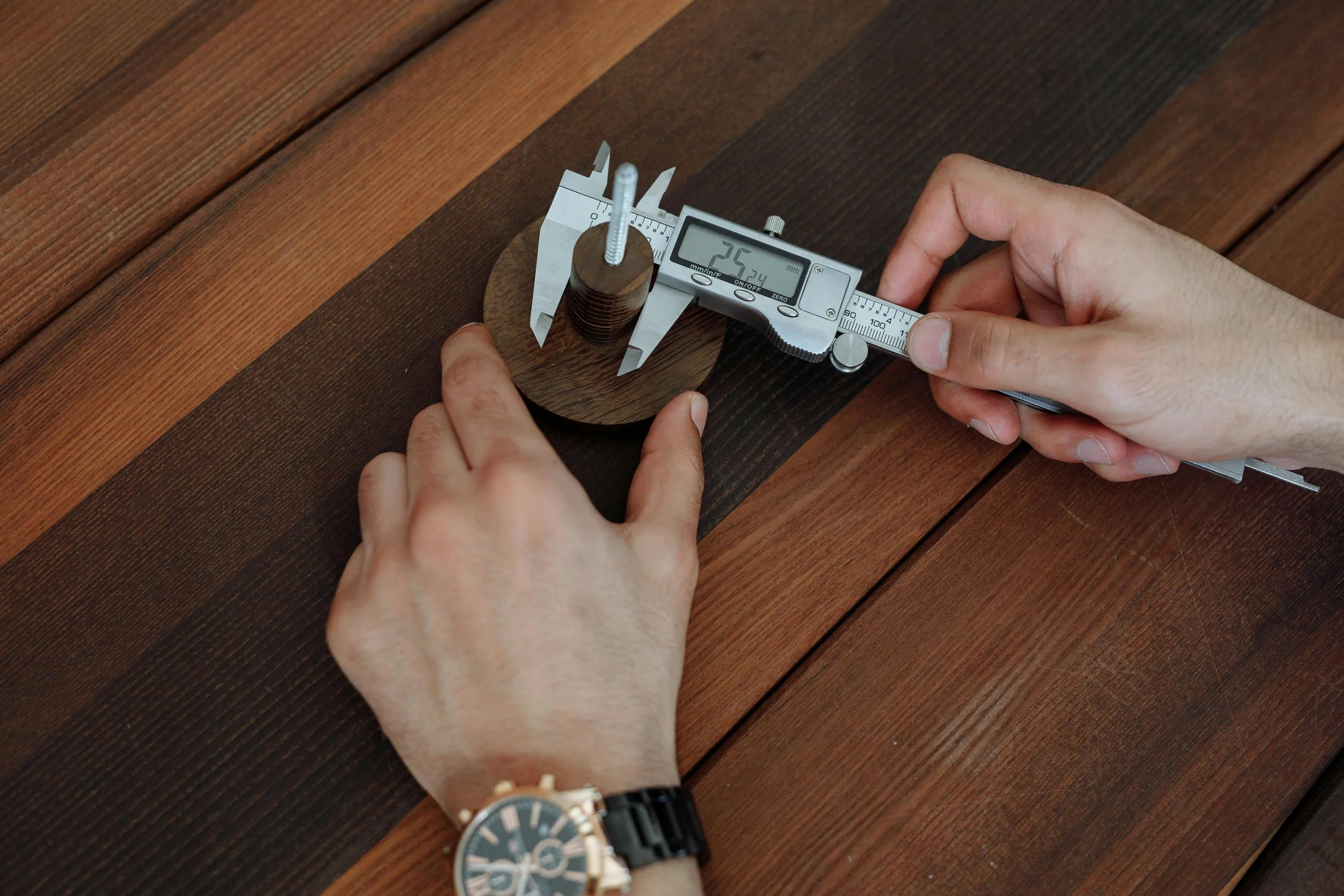 A person measuring the thickness of a wood sample with a digital caliper on a wooden surface.