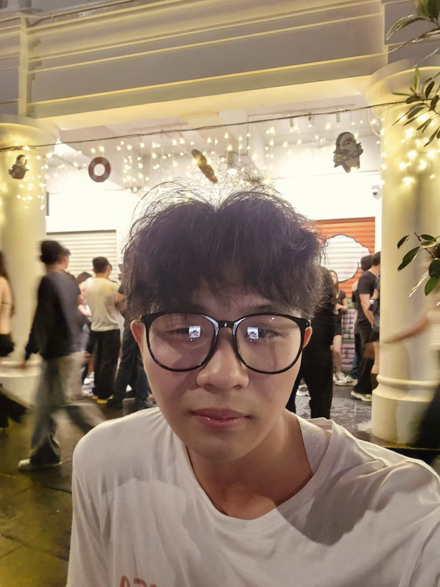 A young man with glasses and curly hair taking a selfie at a crowded indoor event with string lights and nautical decorations in the background.