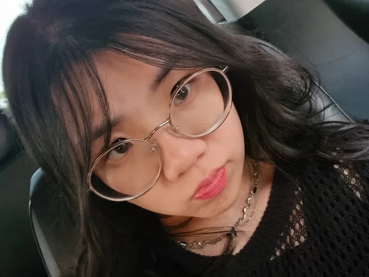 A young woman with long black hair, wearing large round glasses, a black patterned top, and layered necklaces, sitting in a car.