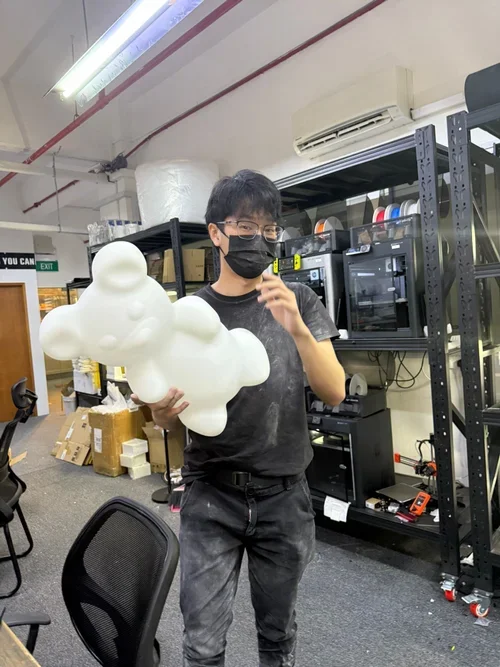Person wearing glasses and a mask holding a white foam cloud-shaped object in a workspace with equipment and boxes.