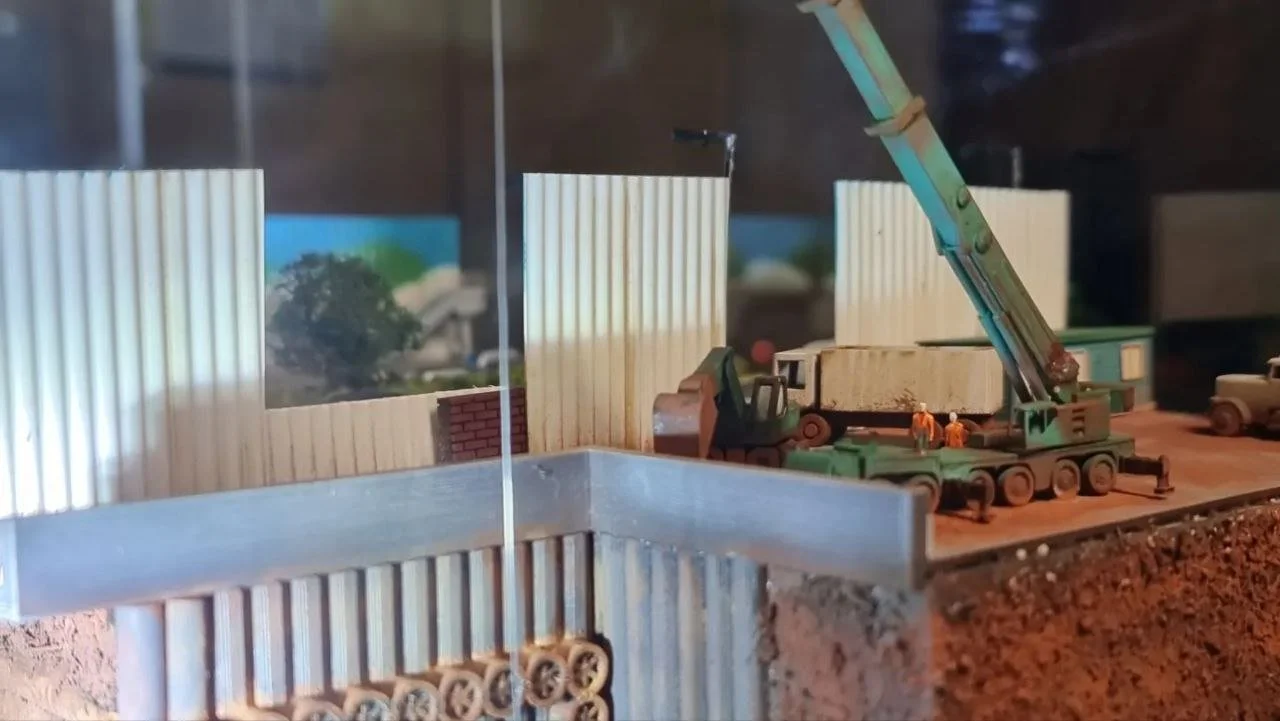 Miniature construction site with a crane, workers, and vehicles, enclosed by a white picket fence.