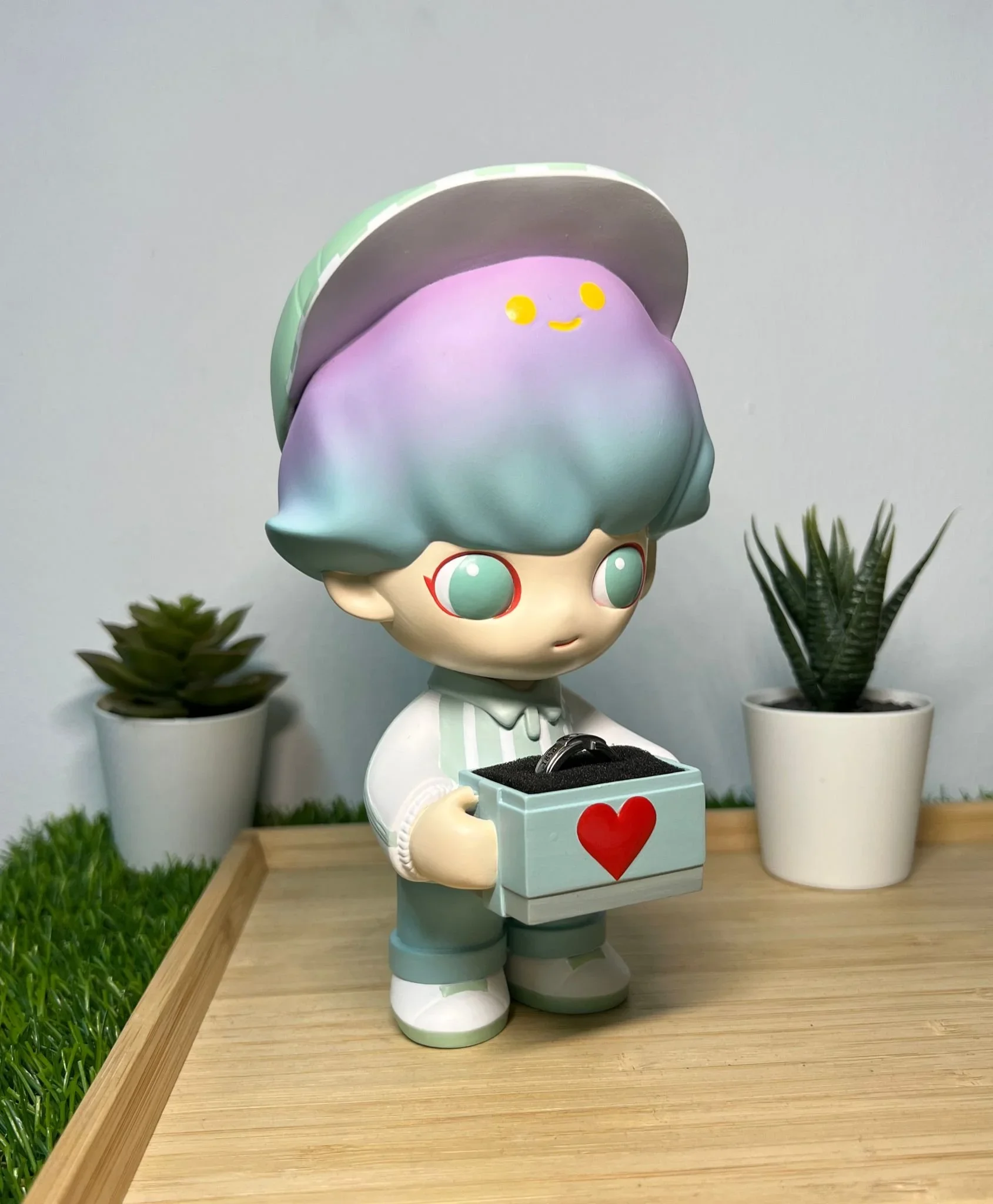 A cute cartoon-style doll with pastel-colored hair holding a gray box with a red heart, standing on a wooden surface with two potted plants in the background.